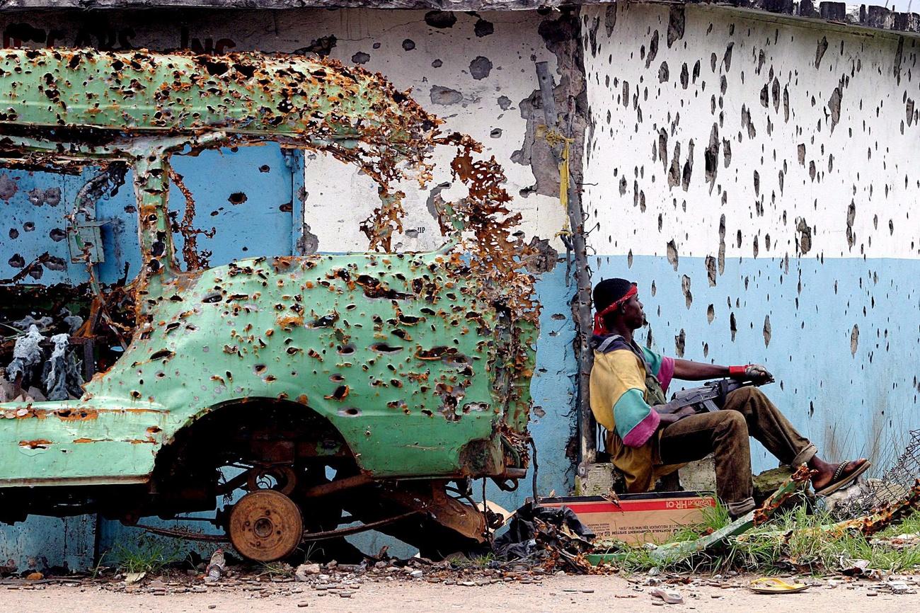 Armed man in Liberia sitting next to a car wreck riddled with bullets