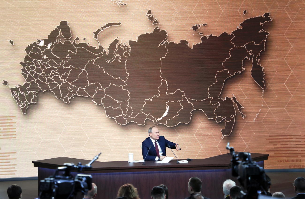 Vladimir Putin seduto al tavolo di una grande sala stampa; dietro di lui, enorme cartina muta della Federazione russa