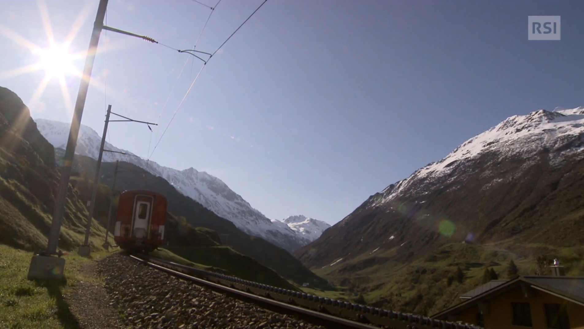 Immagine controsole di una vallata alpina percorsa da binari; un treno si allontana