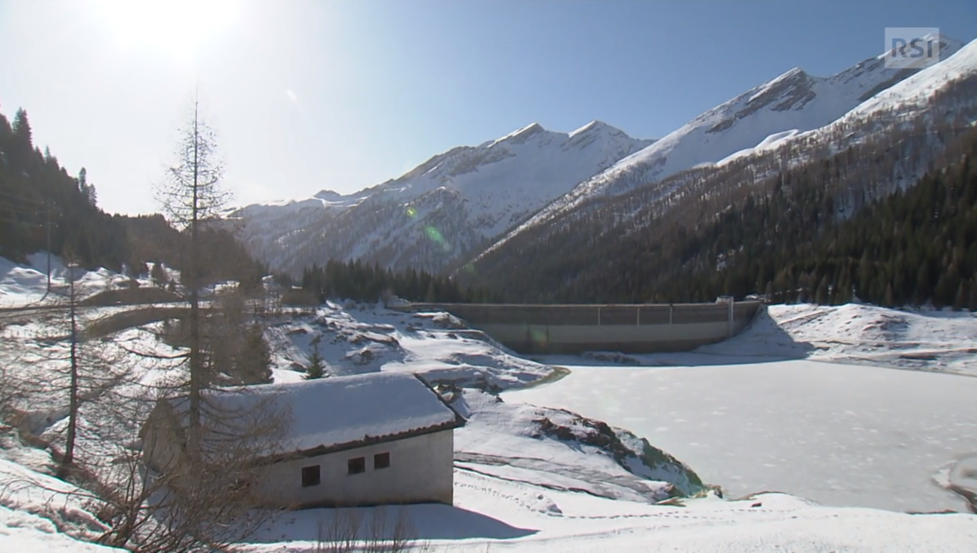 Lago artificiale, con diga di sbarramento ben in vista e superficie completamente ghiacciata; paesaggio alpino innevato