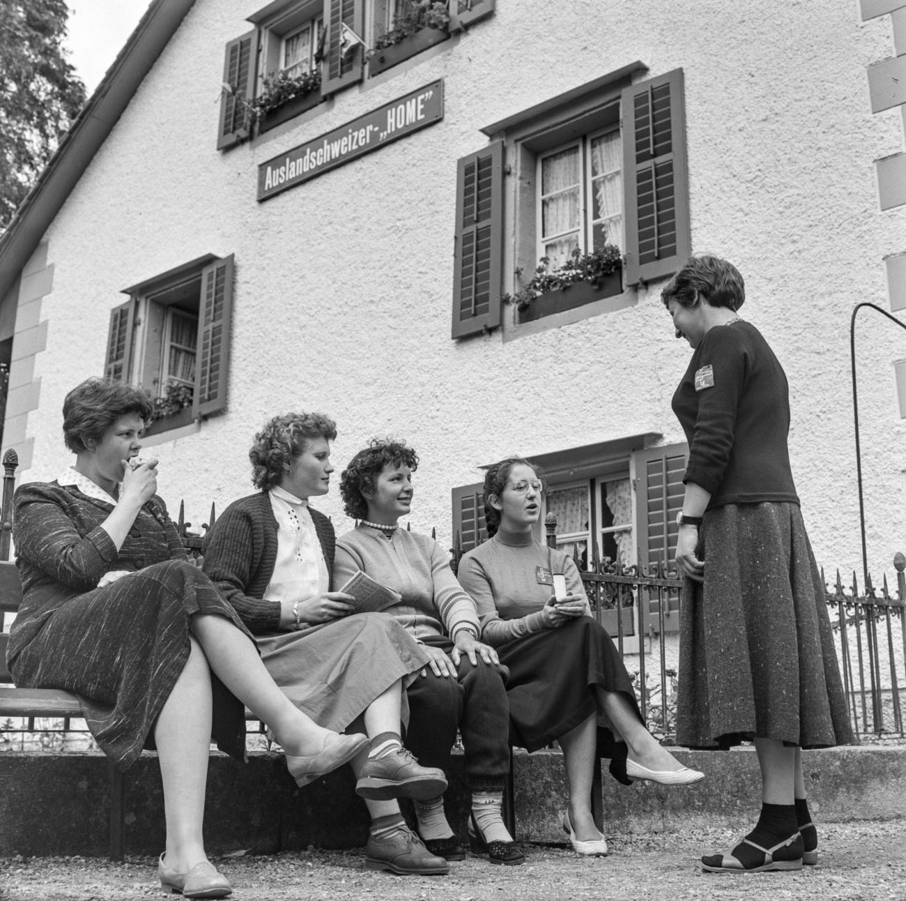 quattro donne sedute su una panchina e una in piedi che chiacchierano, davanti a una casa.