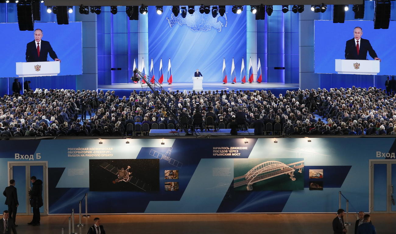 Immagine complessiva di un enorme sala gremita, con maxi-schermi e, lontano, un pulpito da cui parla Putin