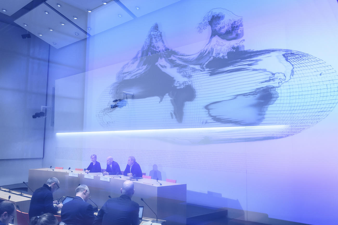 Tre persone sedute a un tavolo di conferenza stampa; alle loro spalle illustrazione di un globo terrestre