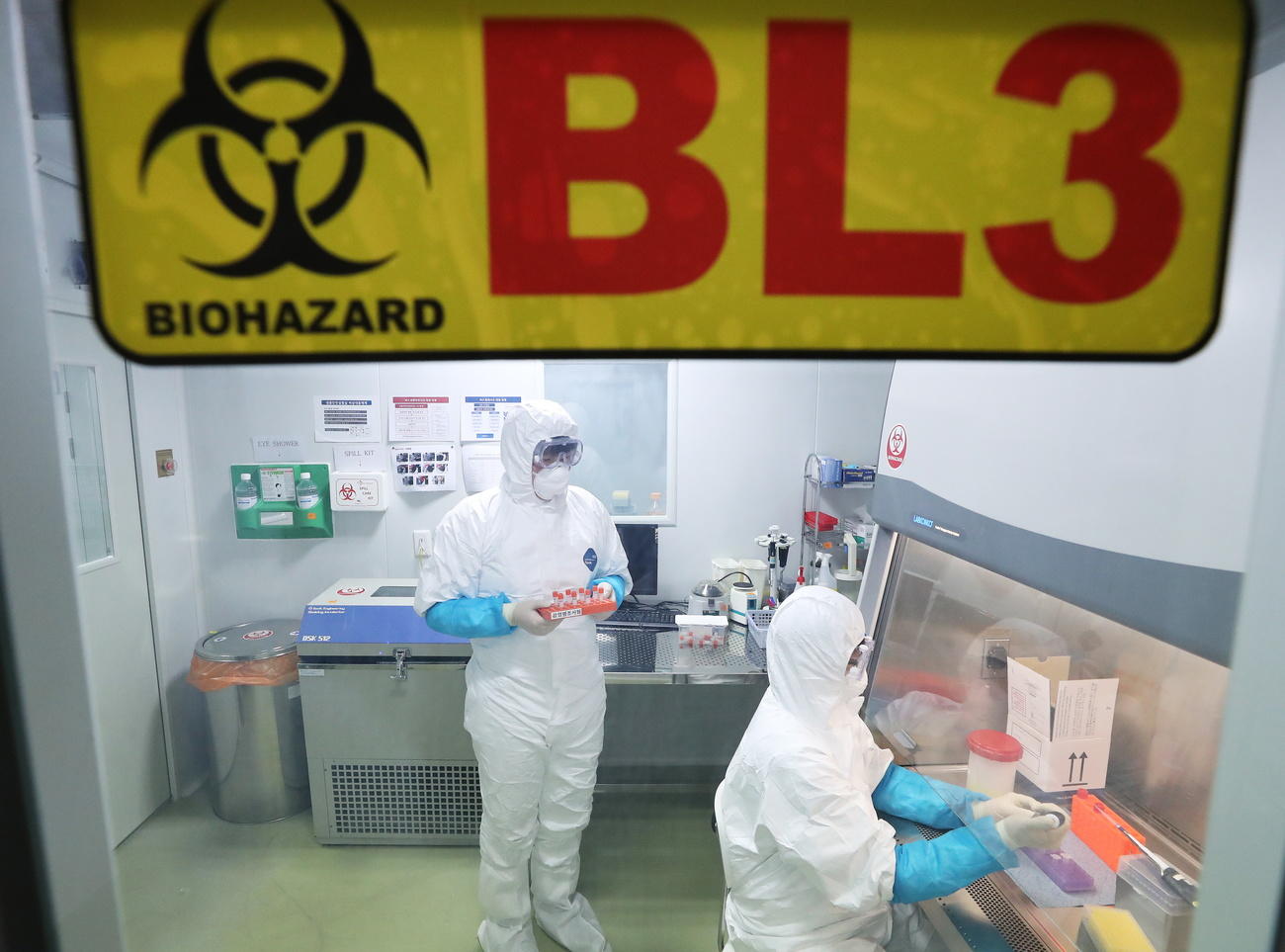 Interno di un laboratorio; operatori completamente coperti, protetti da maschera e guanti; cartello BL3