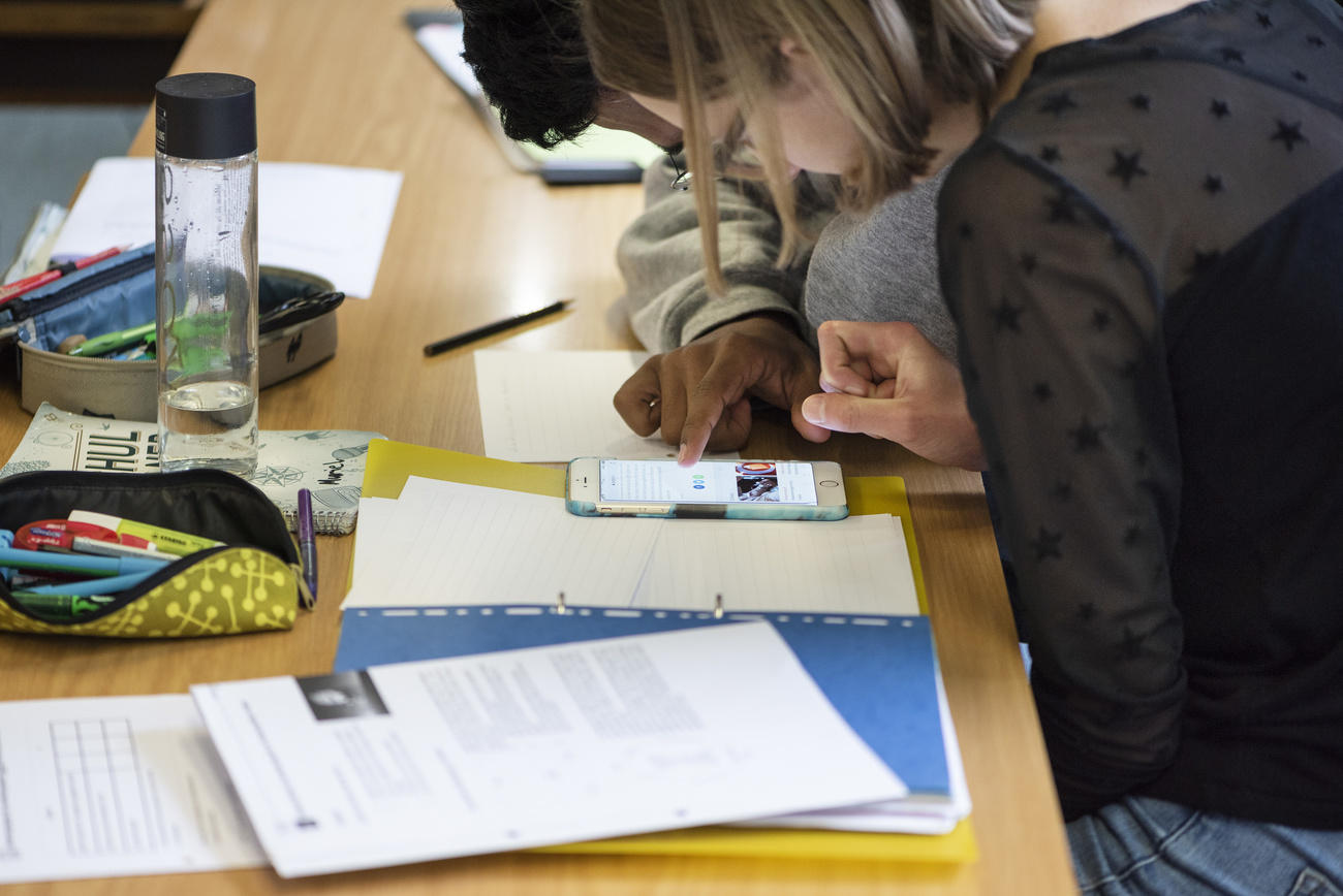 Giovane consulta lo smartphone sui banchi di scuola (si vedono bene apparecchio e astuccio, i volti e l ambiente sono sfocati)