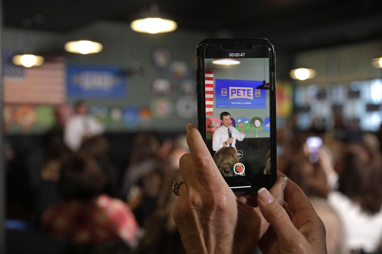 Immagine di Pete Buttigieg con bandiere americane alle sue spalle, ripreso con un telefonino.