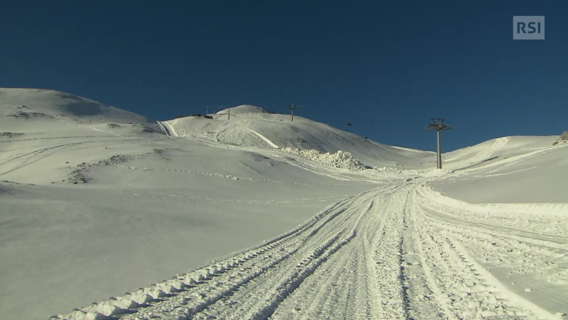 Paesaggio montano innevato con solchi come di passaggio di un veicolo; in lontananza, seggiovia