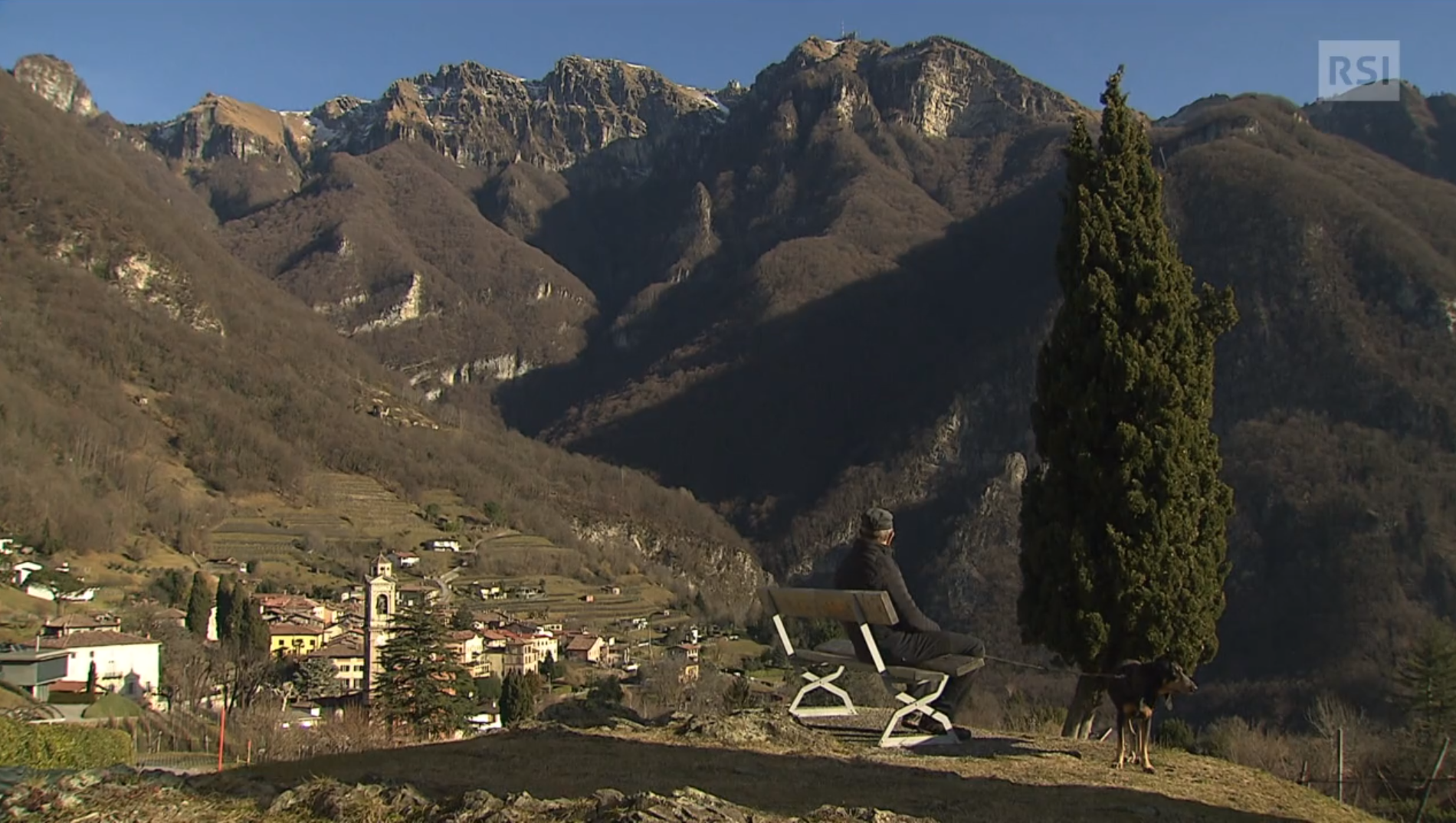 Paesaggio alpino, senza neve; villaggio in basso a sinistra; un anziano con un cane su una panchina guarda panorama