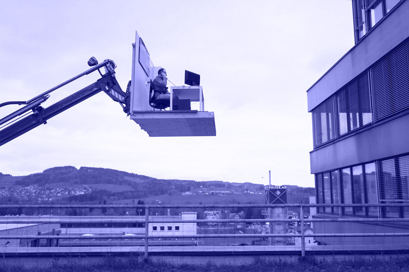 a man sitting at a office desk. The desk is placed at the fork of a tractor outside the window of a office building