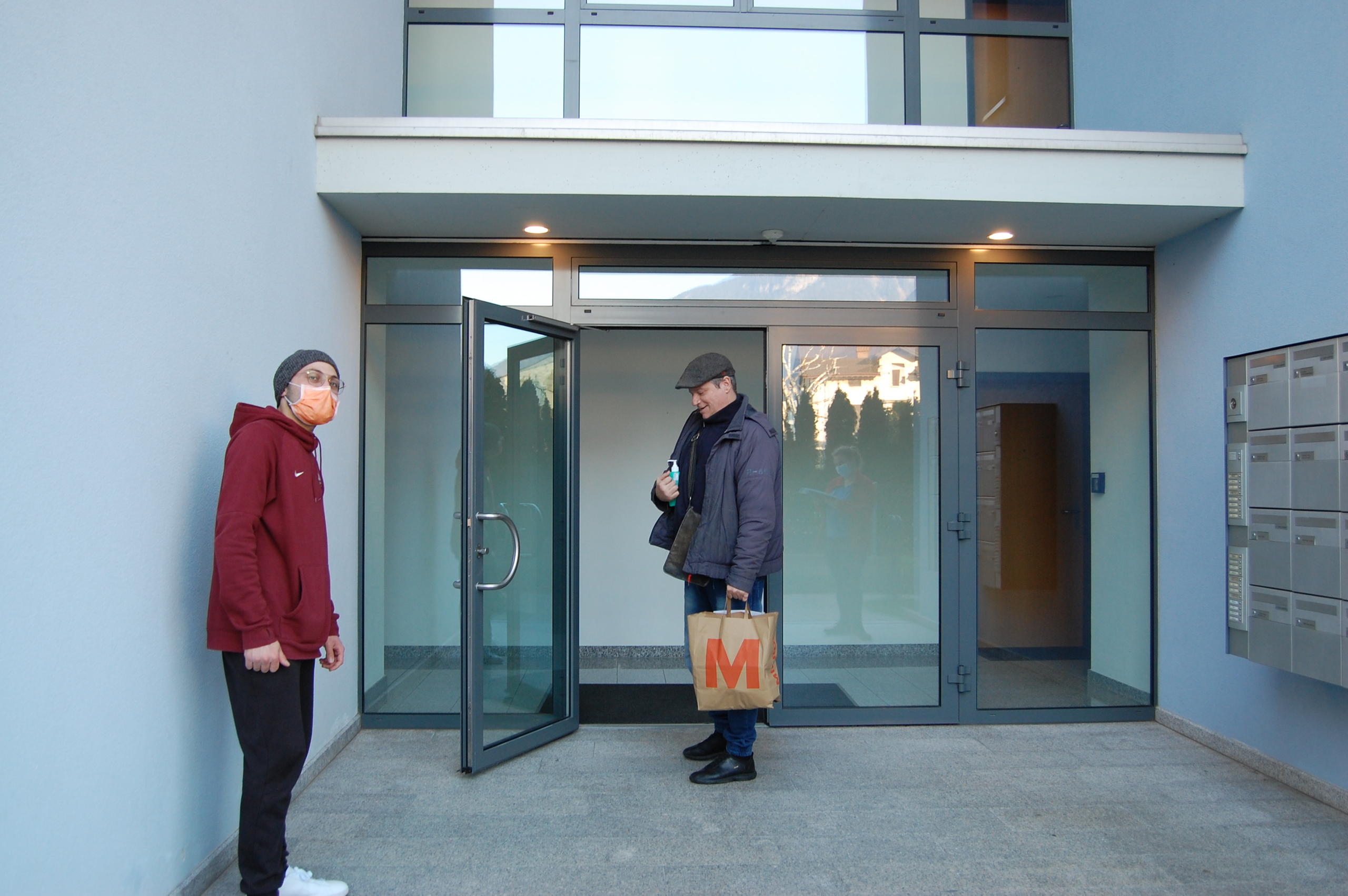 Un uomo con una sporta in mano all ingresso di un edificio e un giovane che resta a distanza