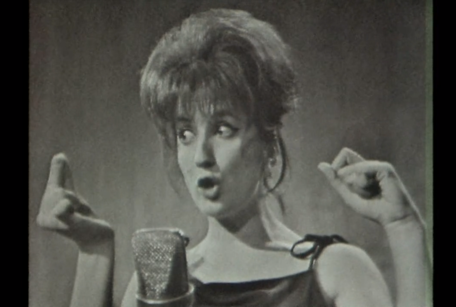 (b/n) Primo piano di una donna che canta davanti a un microfono da studio; fa un gesto con la mano; sfondo neutro