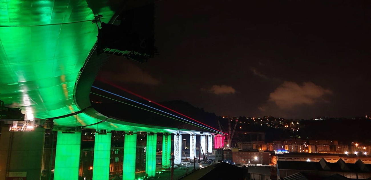 Immagine di un lungo viadotto illuminato dal basso con luci verdi, bianche e rosse; attorno, città di notte.