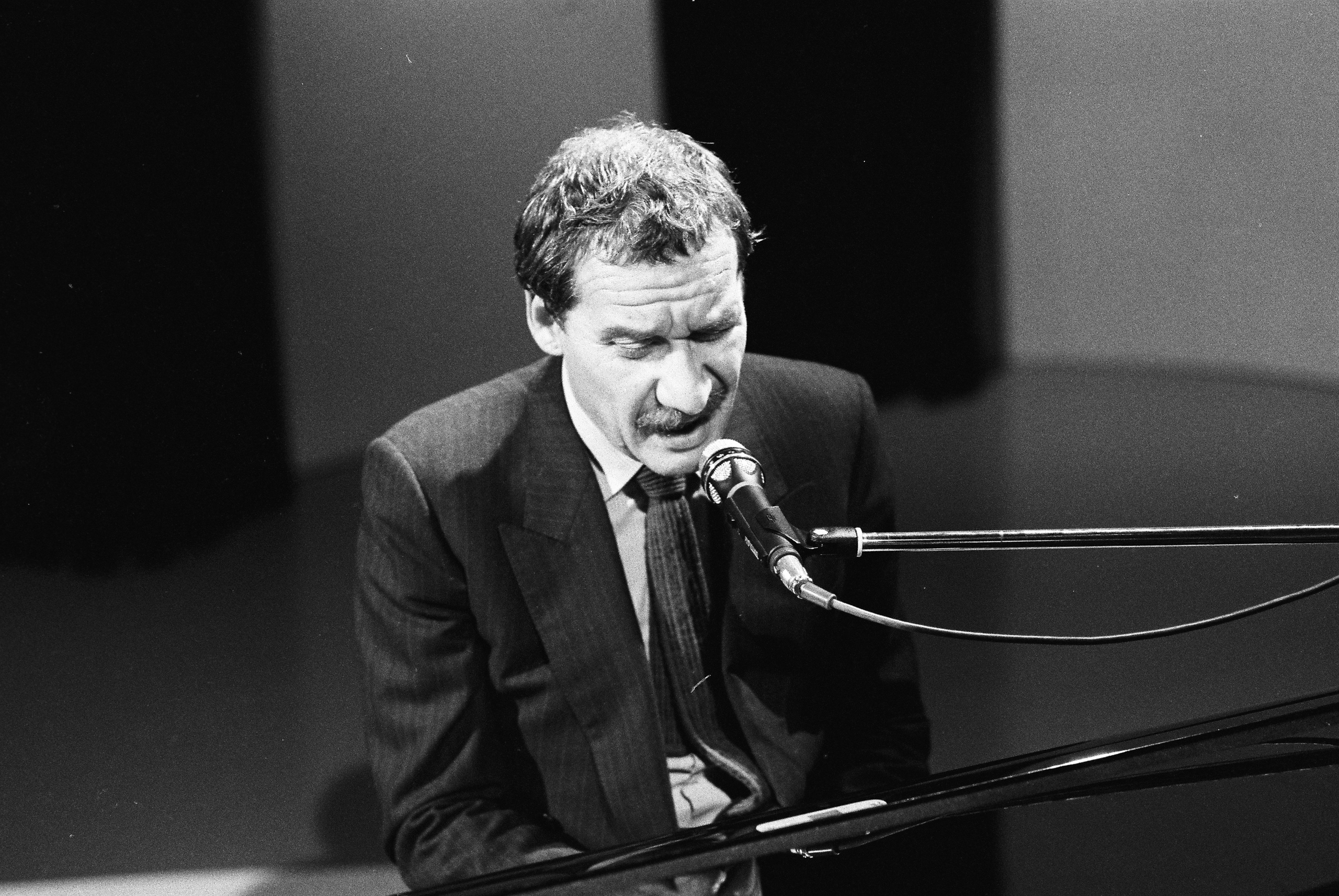Un uomo in giacca e cravatta seduto al pianoforte, suona e canta in un teatro o studio televisivo.