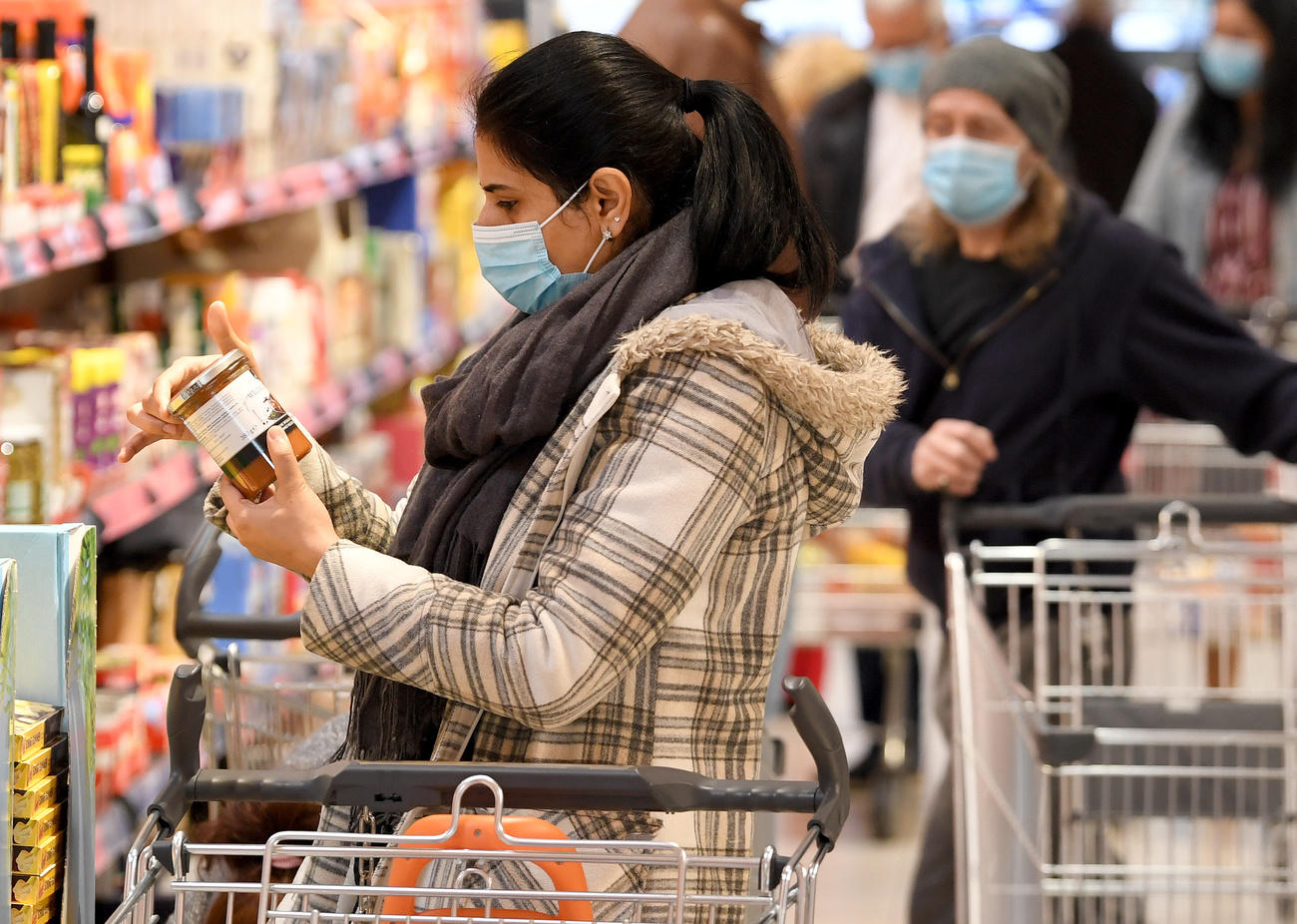 Corsia di un supermercato. Una dona scruta un vasetto, un altra persona spinge un carrello, entrambi indossano mascherina