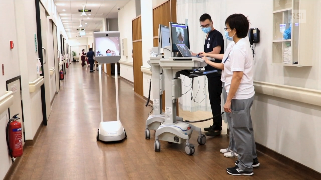In un corridoio d ospedale, carrello con schermo sul quale appare il volto di un infermiera. Personale lo telecomanda.