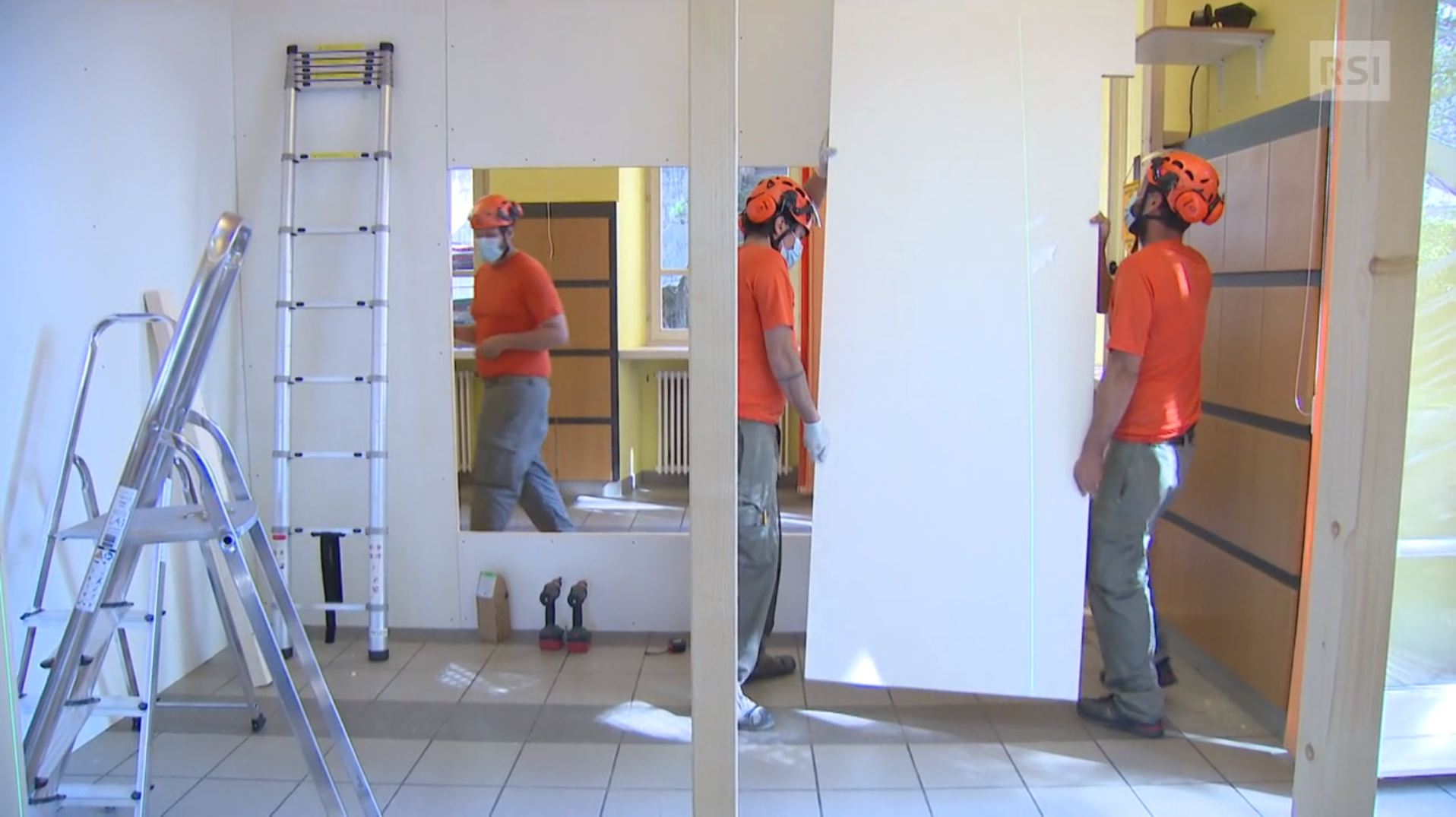 Tre uomini in abiti da lavoro, con maglietta arancione, montano pannelli di separazione in una stanza.