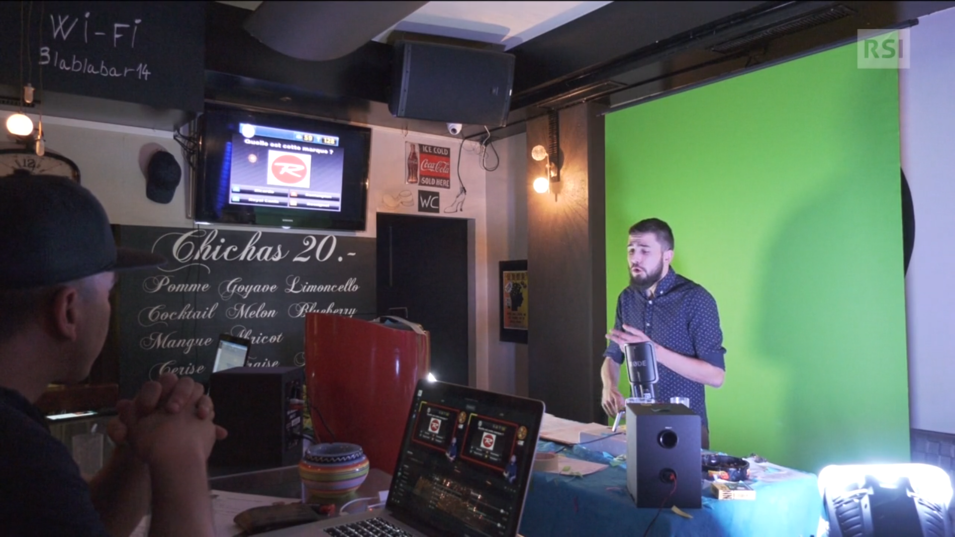 Giovane uomo parla davanti a un microfono verso una telecamera all interno di un piccolo bar; sugli schermi attorno domande quiz