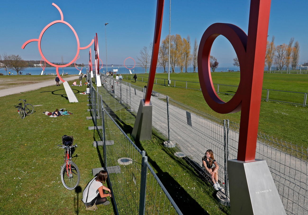 Un parco vicino al lago con un alta barriera metallica lungo un sentiero sterrato in grande prato; persone sui due lati