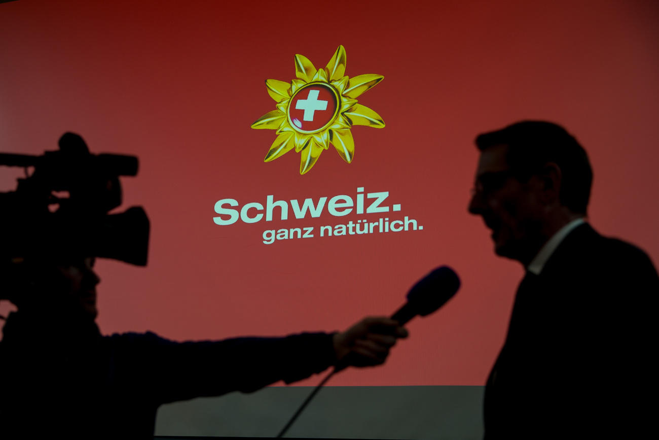 Silhouette di un telegiornalista e di un uomo che viene intervistato; a fuoco il fondo rosso col logo di Svizzera Turismo