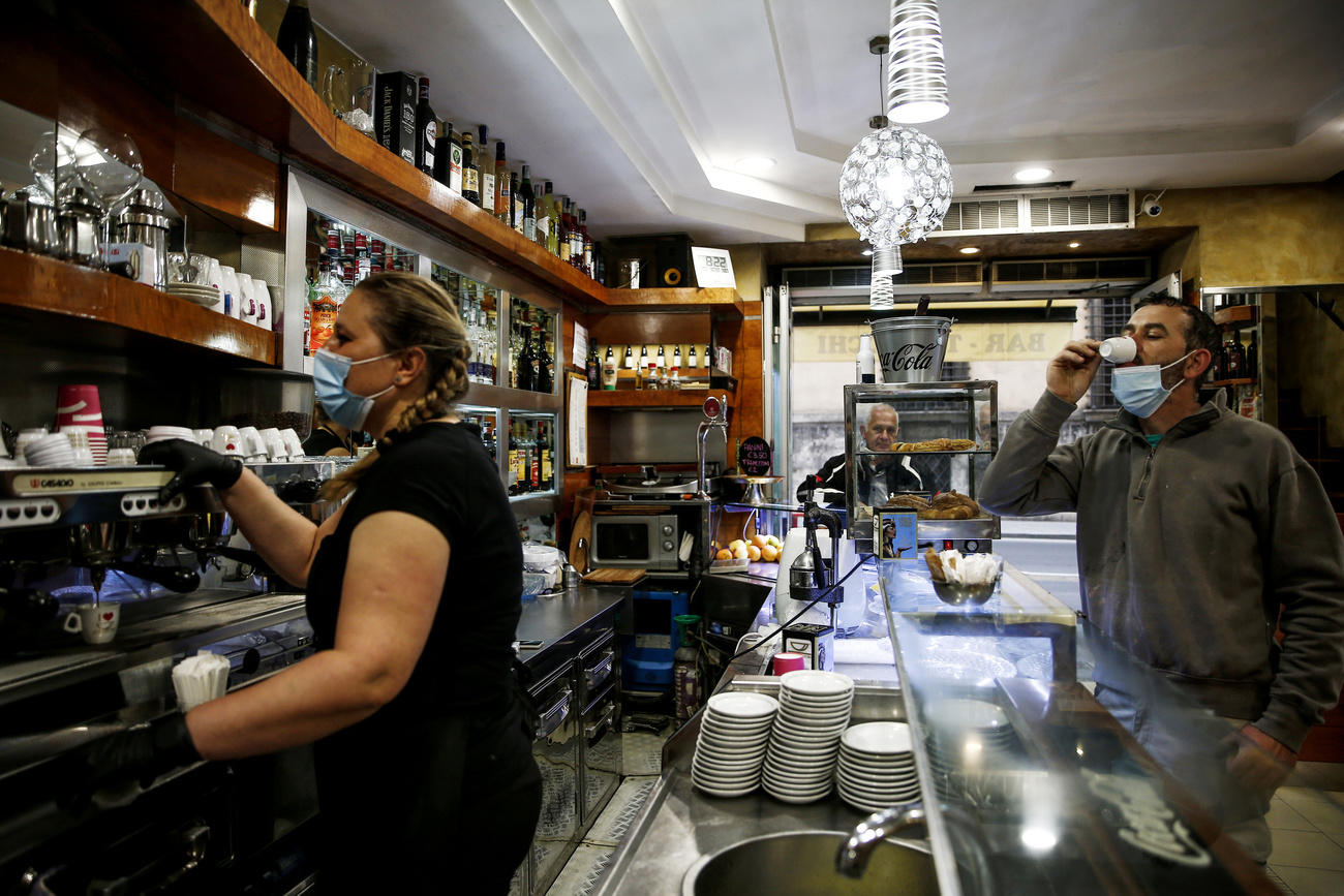 Interno di un bar; barista prepara un caffè, avventore ne beve uno; tutti indossano la mascherina.