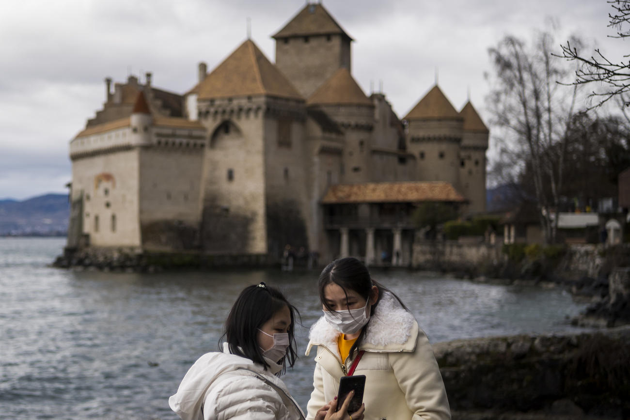 Un castello medievale in riva a un lago visto da 50 metri, in primo piano due turisti asiatici con mascherina