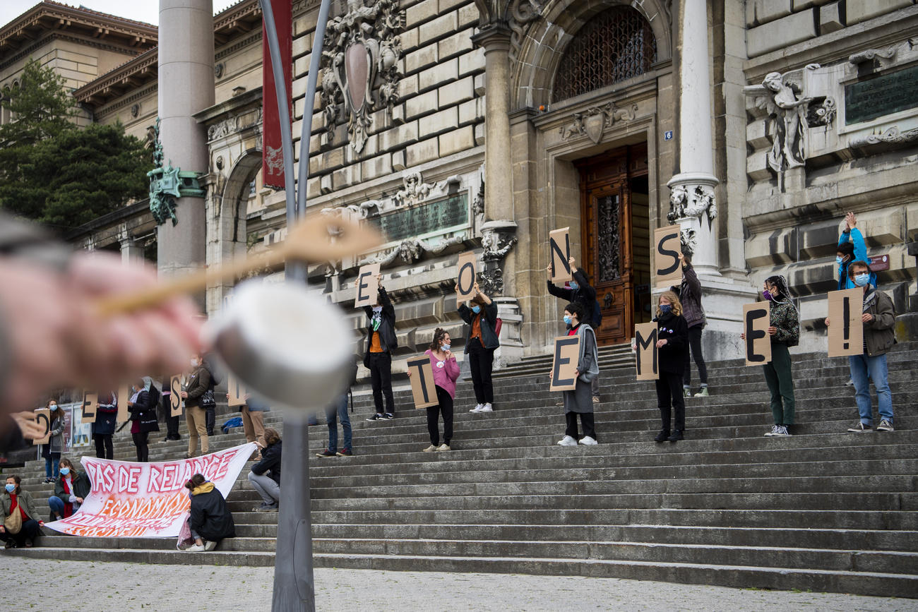 Scalinata di un vecchio edificio con giovani che reggono ognuno una lettera di uno slogan; in prim piano, mestolo e padella