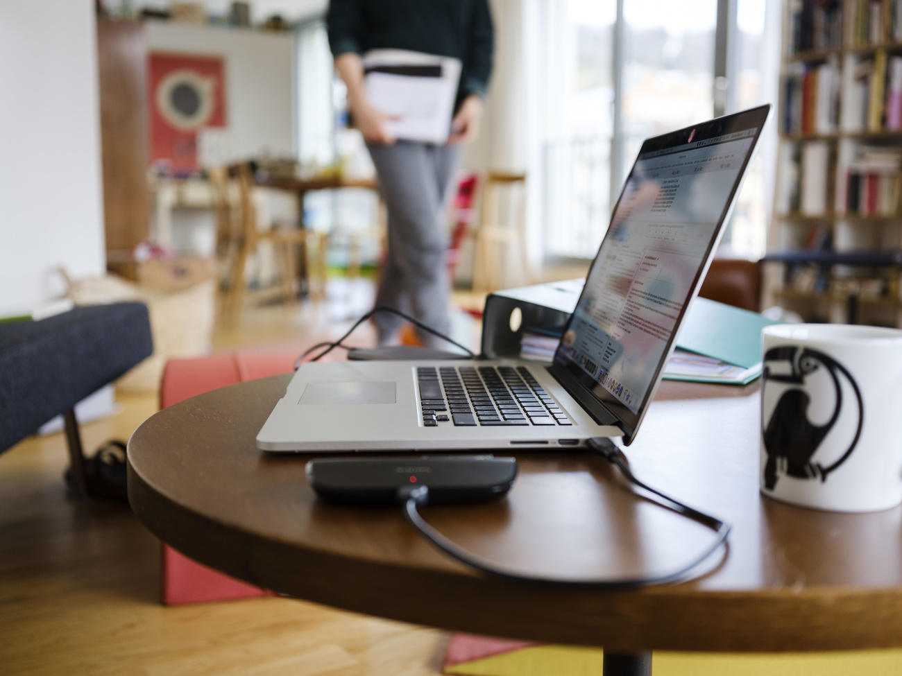 Tavolino da soggiorno con computer, classificatore e tazza posati. Si intravvedono persona, libreria, giocattoli