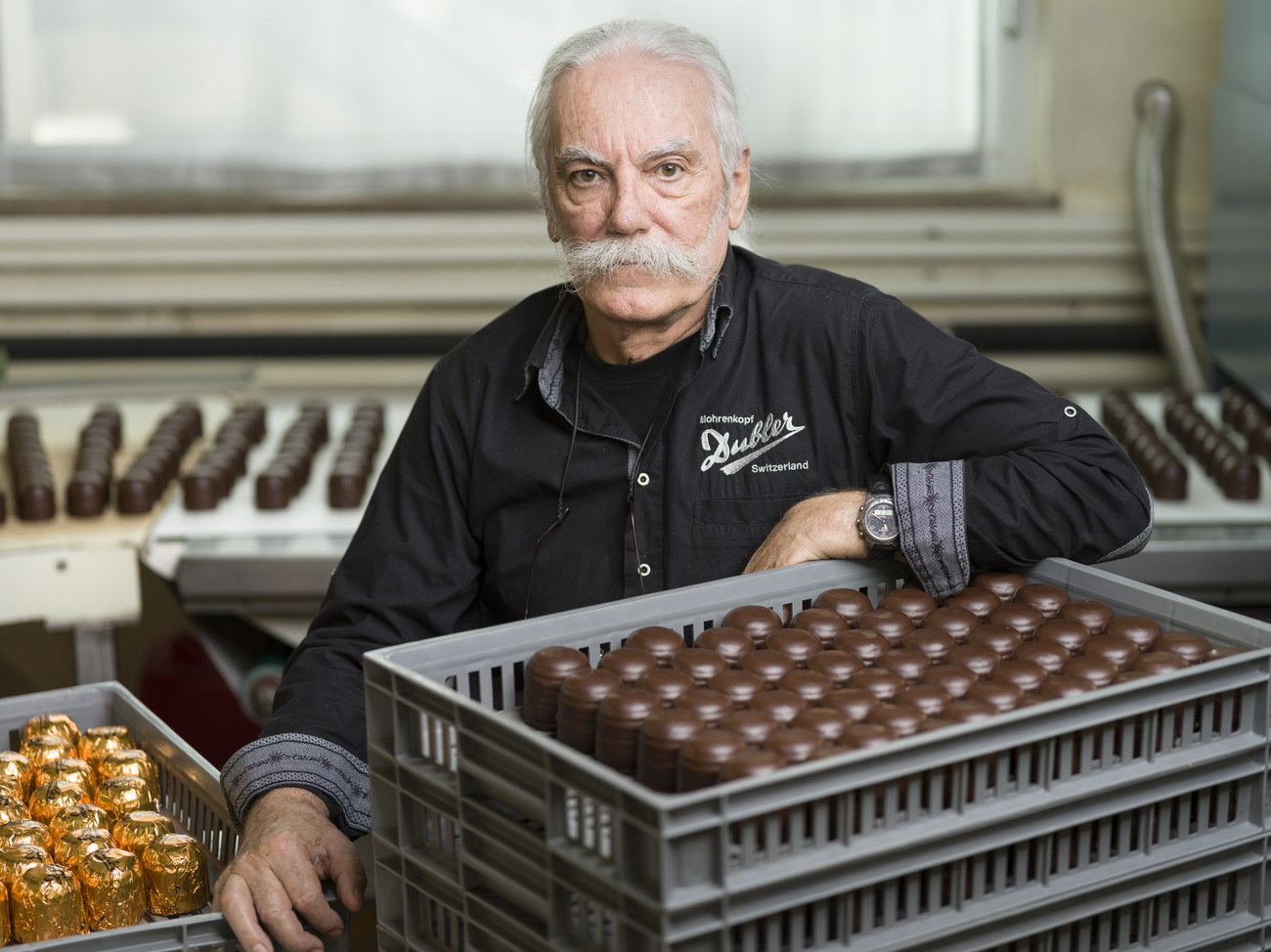 un uomo con una tuta da lavoro seduto in una fabbrica davanti a una cesta piena di moretti, dolciumi tipici svizzeri.