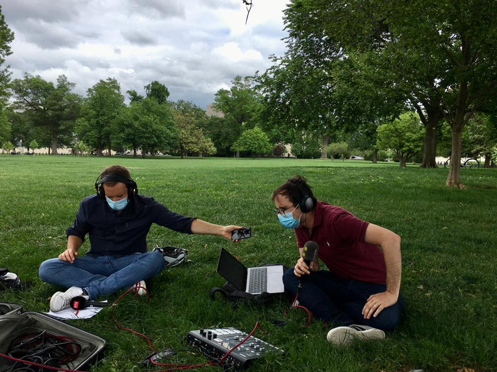 Zwei Männer sitzen mit Masken und Mikrofonen auf einer grünen Wiese am Boden.