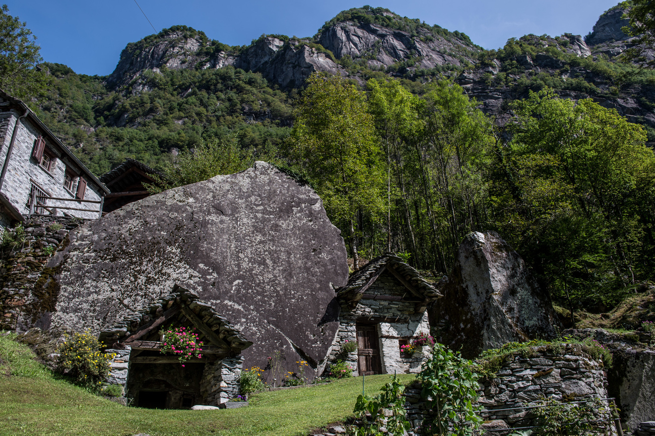 Unter einem Felsblock am Fuss eines Steilhangs erbautes Steinhaus mit Wald im Hintergrund