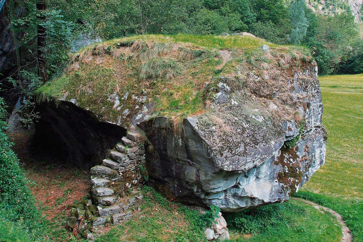 Felsbrocken am Rand eines Waldes mit Grad darauf und Wiese darunter und einer steinernen Treppe, um hinaufzusteigen.