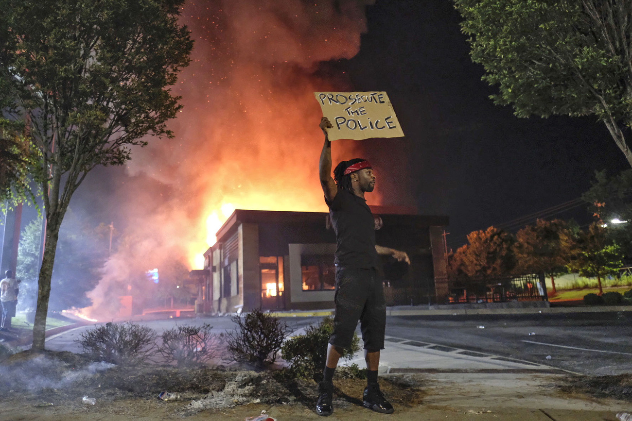 Un uomo di colore davanti al fast food in fiamme con un cartello: Perseguite la polizia