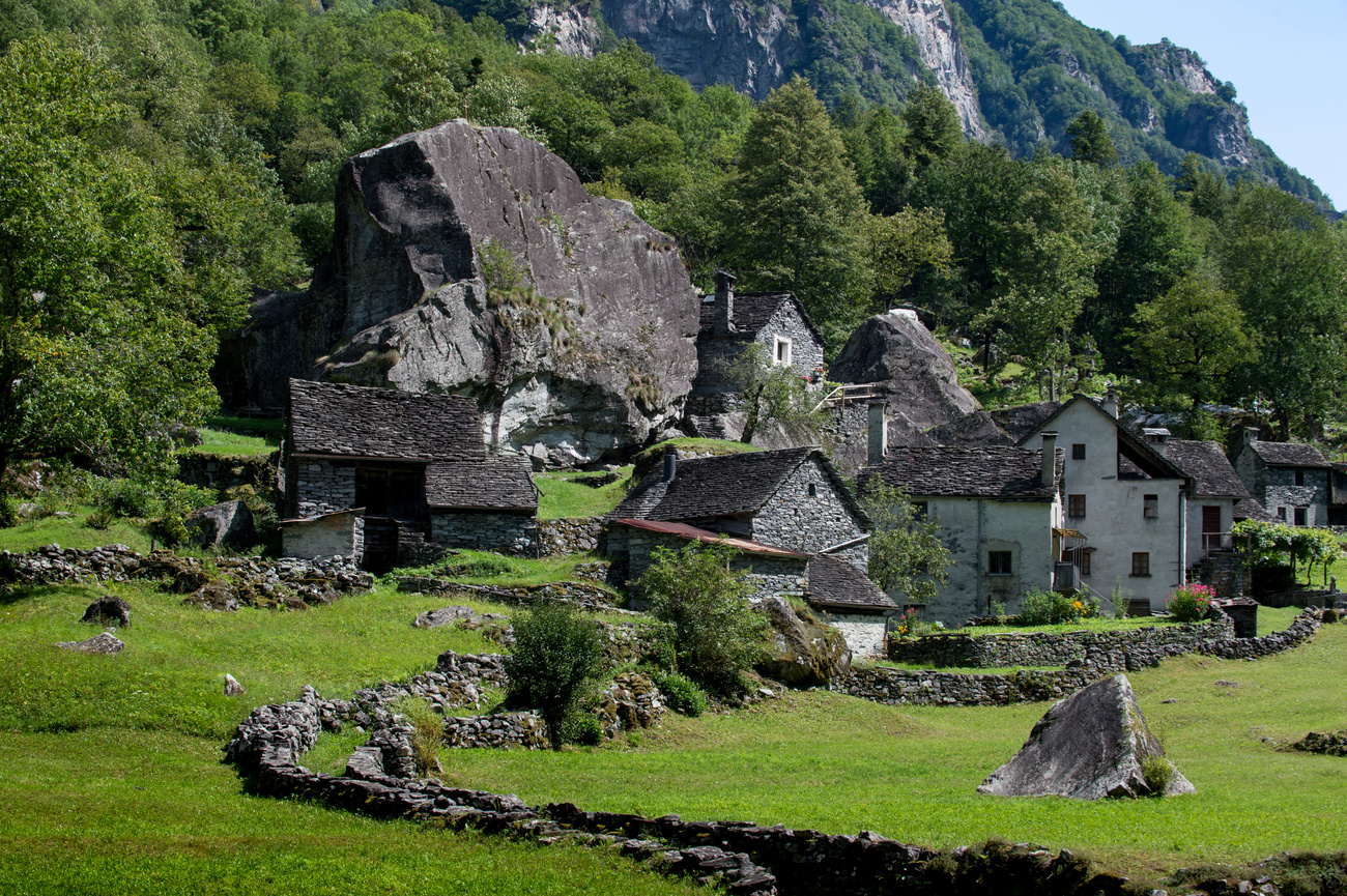 Dorf mit Steinhäusern, die neben einem riesigen Felsblock am Fuss eines bewaldeten Hangs gebaut wurden; davor Wiesen