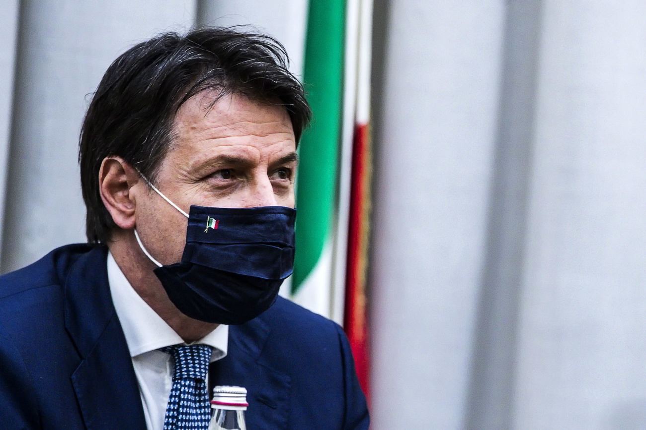 Uomo in abito formale e volto coperto da mascherina nera con bandierina italiana; bandiera grande alle sue spalle