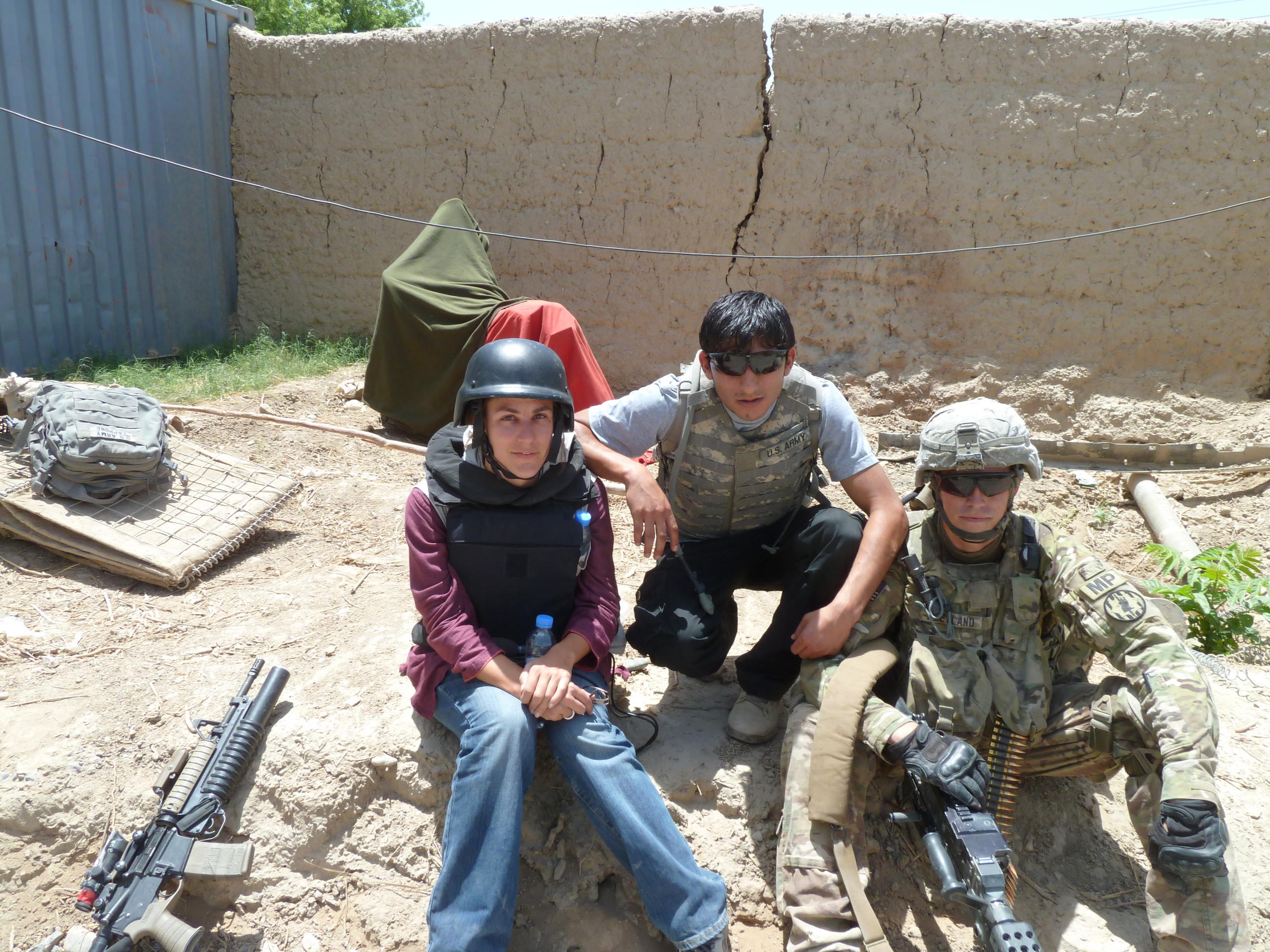 una donna seduta di fianco a due soldati in un accampamento militari. intorno a loro ci sono varie armi.