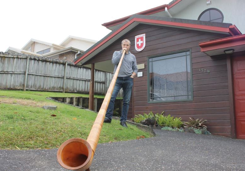Ein Mann steht vor einem Haus und bläst in ein Alphorn.