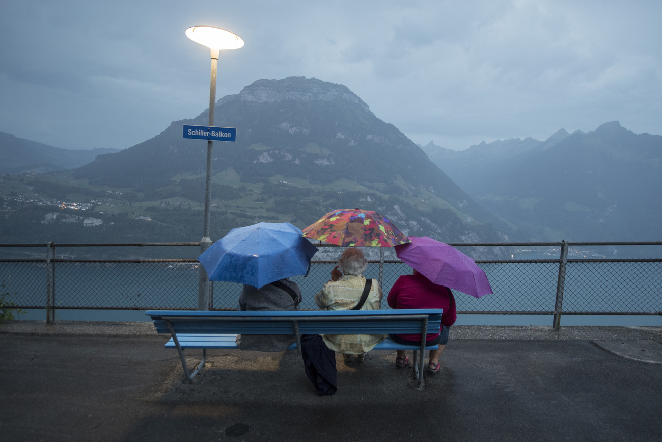Personas mayores sentadas en una banca, con paraguas, frente al lago en tarde lluviosa.