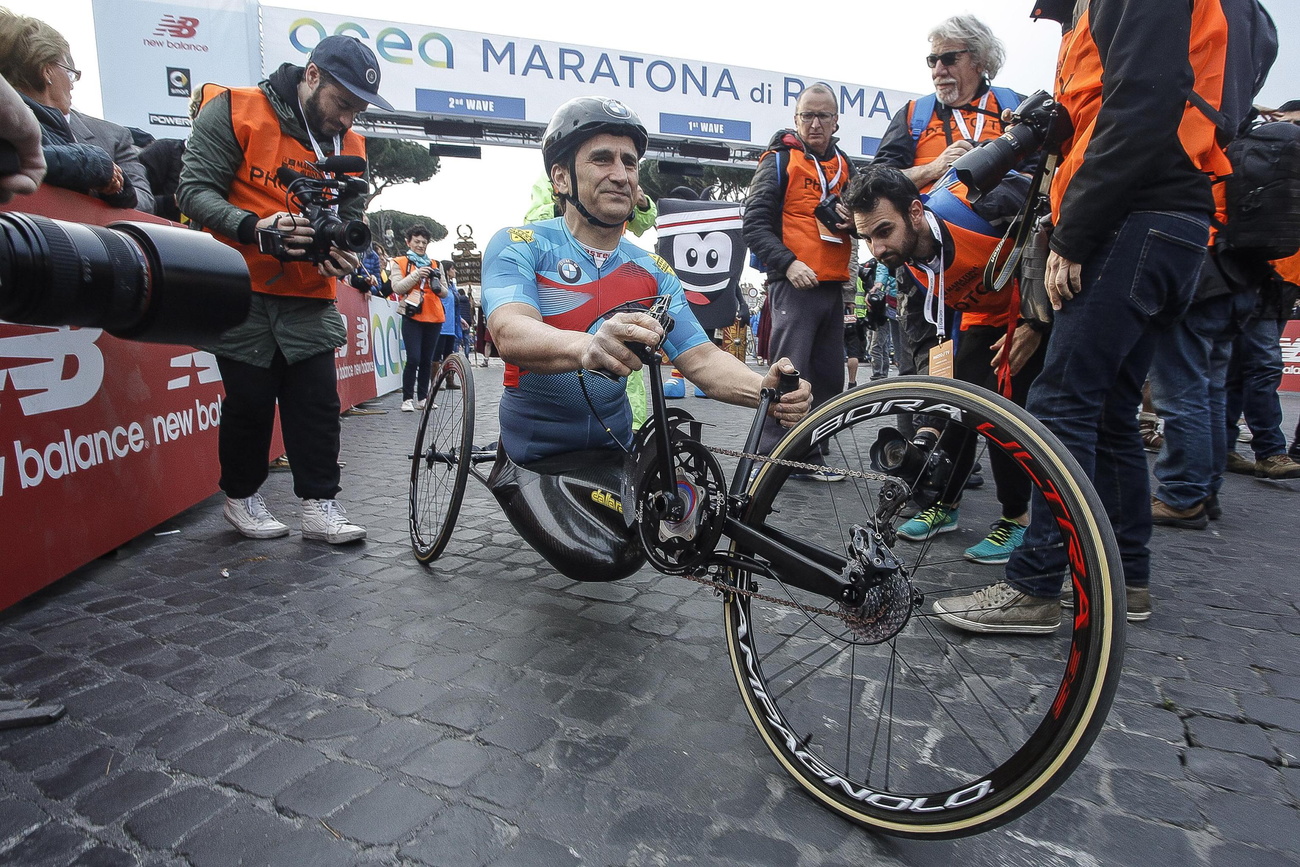 Uomo in tenuta sportiva e in handbike, su strada lastricata di sanpietrini, circondato da fotografi e giornalisti