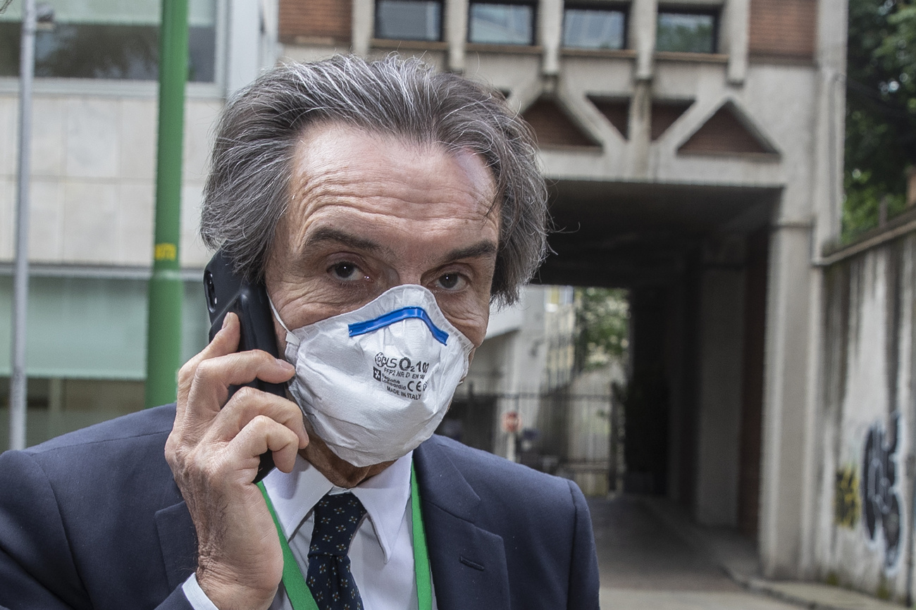 Primo piano di uomo con i capelli bianchi, in abito formale e con la mascherina, parla al telefono.