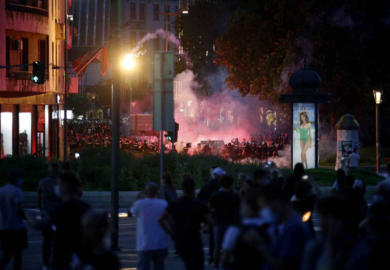 Immagine di centro città con incrocio, semafori, pubblicità. Si intravvedono agenti anti-sommossa manifestanti e fumogeni