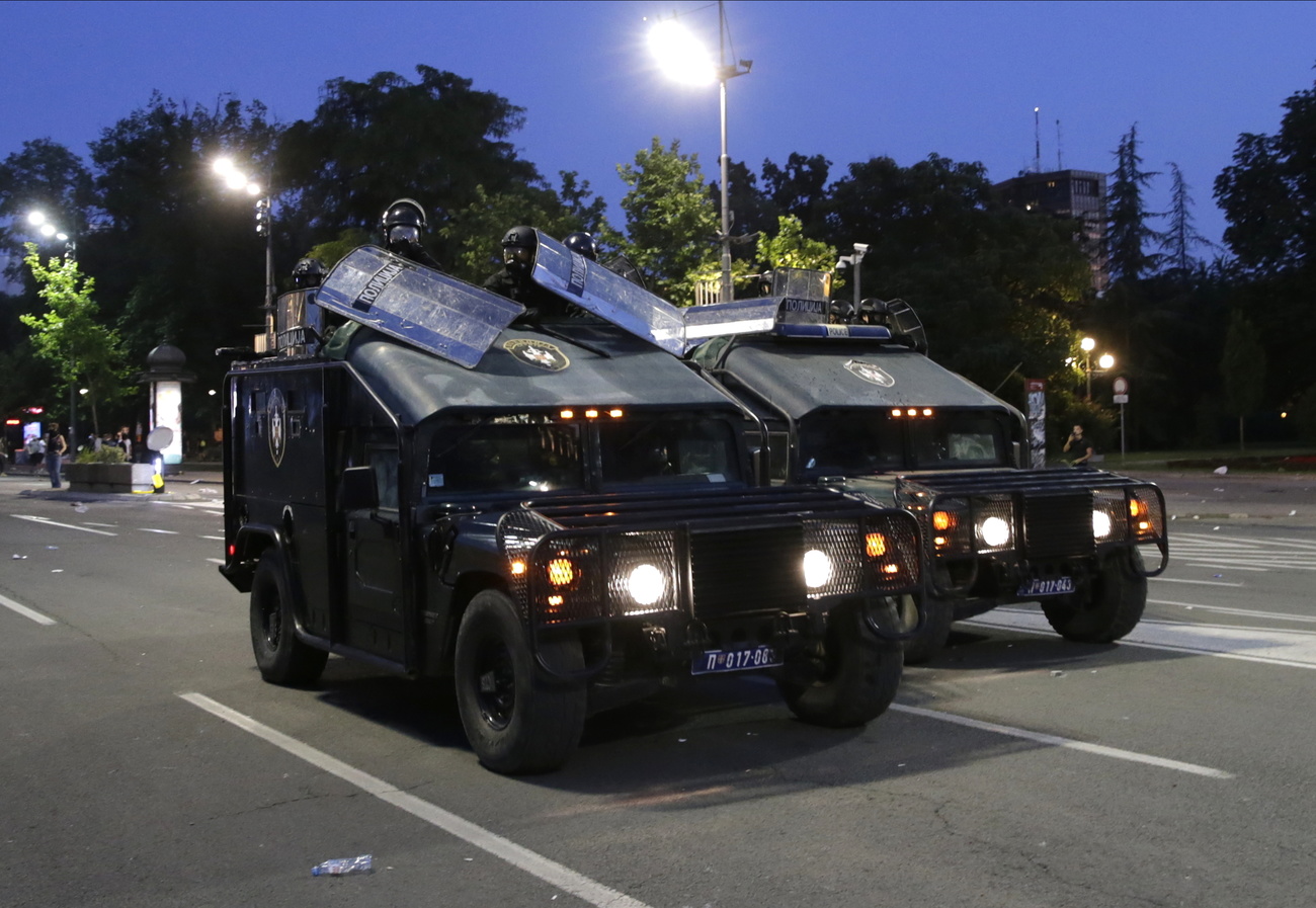 Due hammer e jeep con a bordo poliziotti muniti di scudi su un ampio viale cittadino al crepuscolo; fari accesi