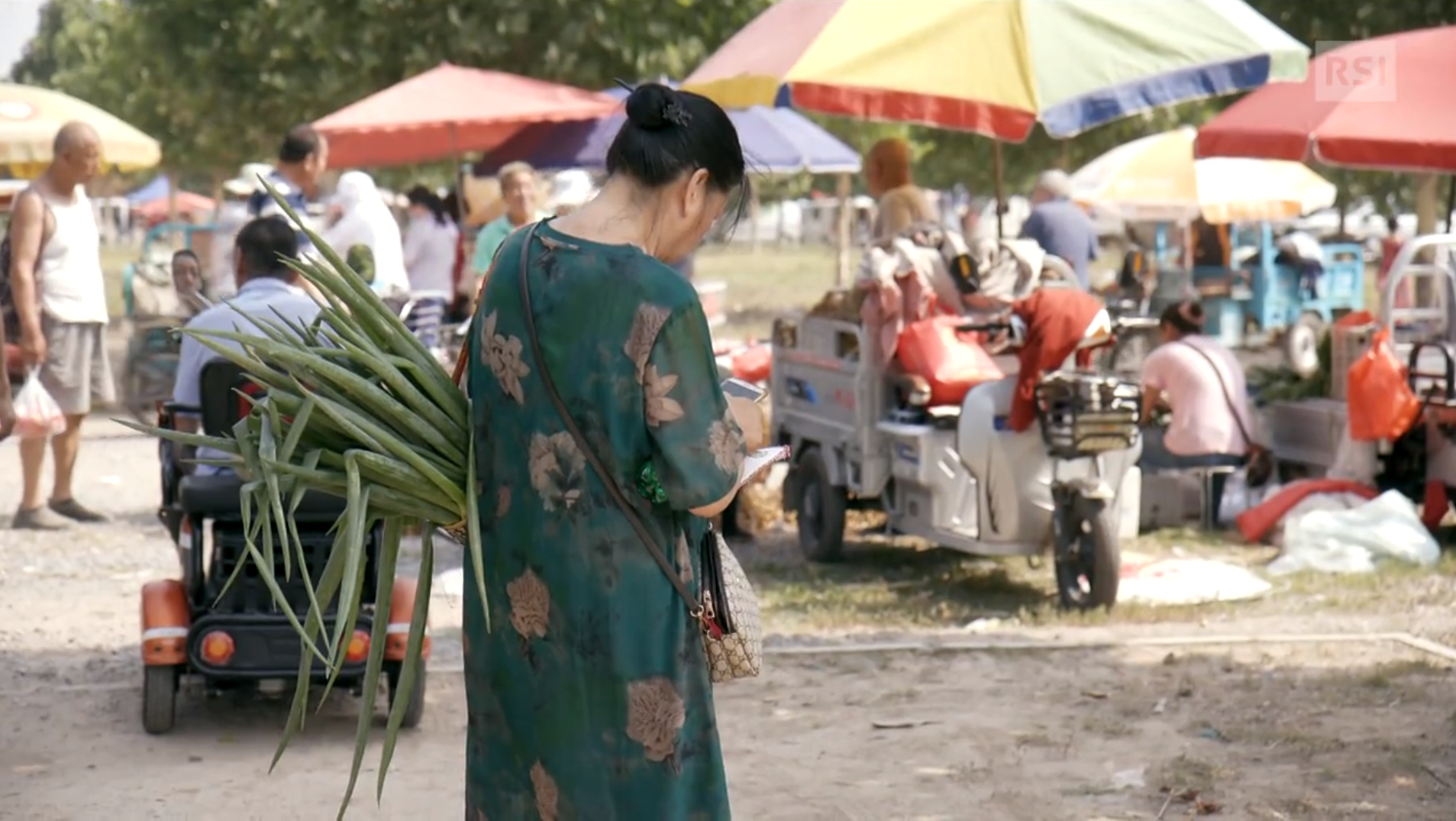 Donna di schiena (con sottobraccio delle verdure) su un piano sterrato con tricicli a motore e ombrelloni
