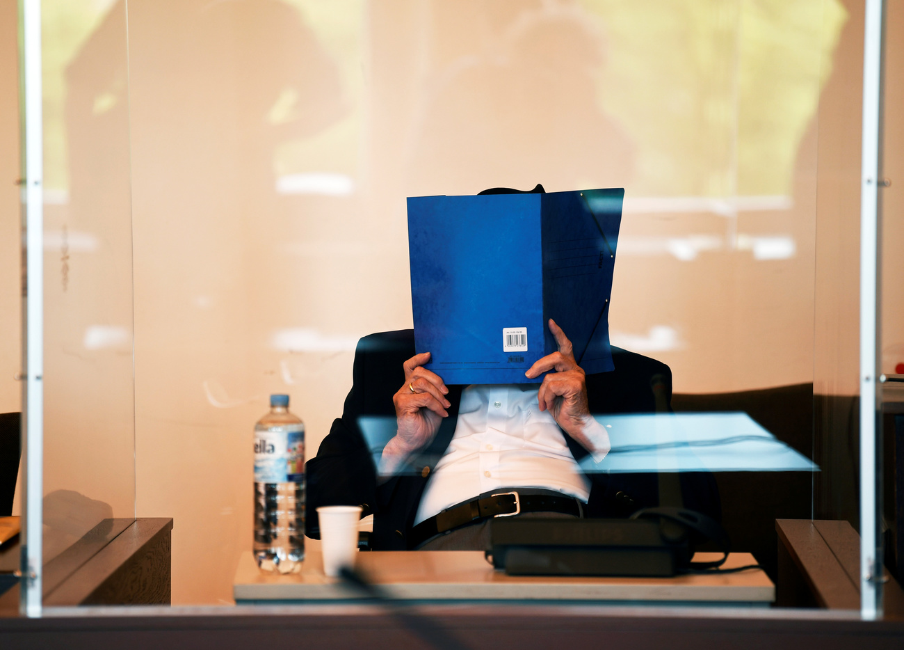 Un uomo seduto a un banco d aula di tribunale, dietro parete di plexiglas, si copre il volto con cartellina