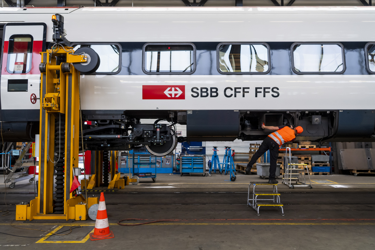 Un vagone ferroviario sollevato in alto in un officina; un operaio, sotto, effettua manutenzione