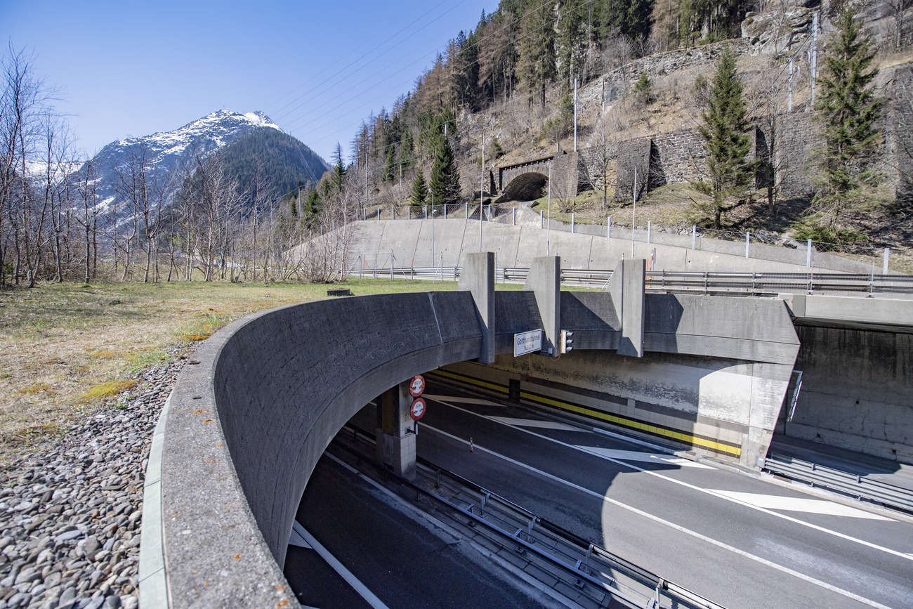 Autostrada imbocca un tunnel prima di un massiccio montuoso; sopra il portale, prati e boschi