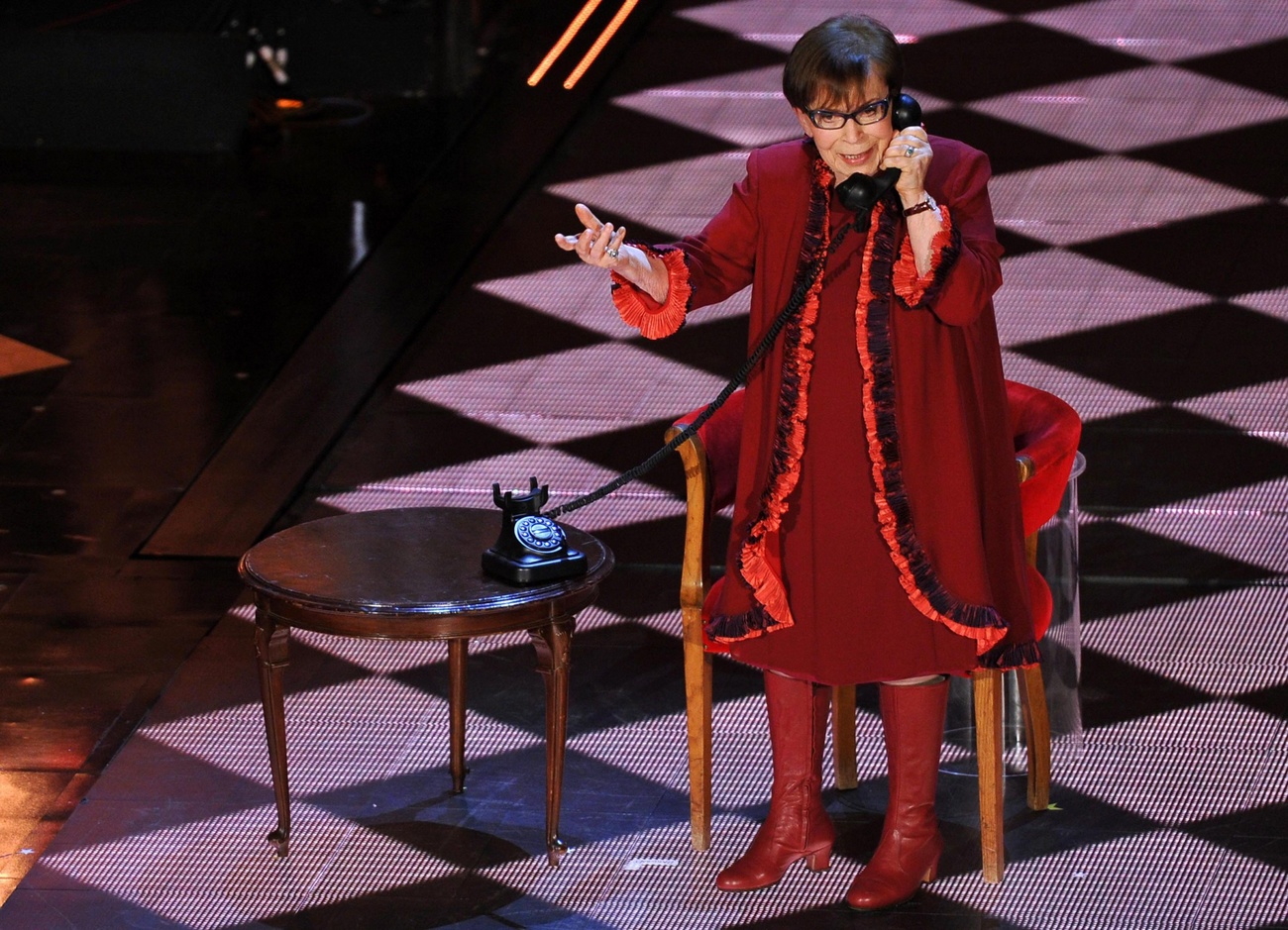 Dona anziana in abito rosso e stivali alti su palcoscenico illuminato a rombi, parla a un vecchio telefono seduta a un tavolino