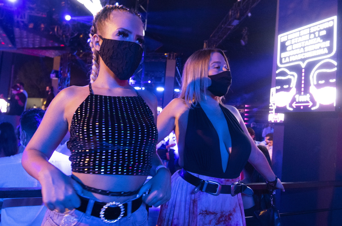 Primo piano di due ragazze in abito da sera in discoteca all aperto; accanto, cartello con indicazioni di misure anti-Covid