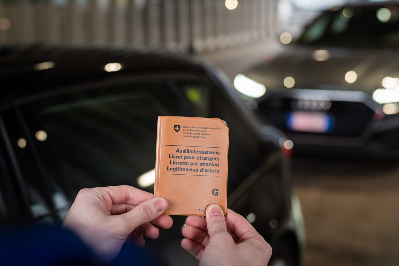 Primo piano di un permesso per stranieri G (astuccio). Si intravvedono auto in coda.