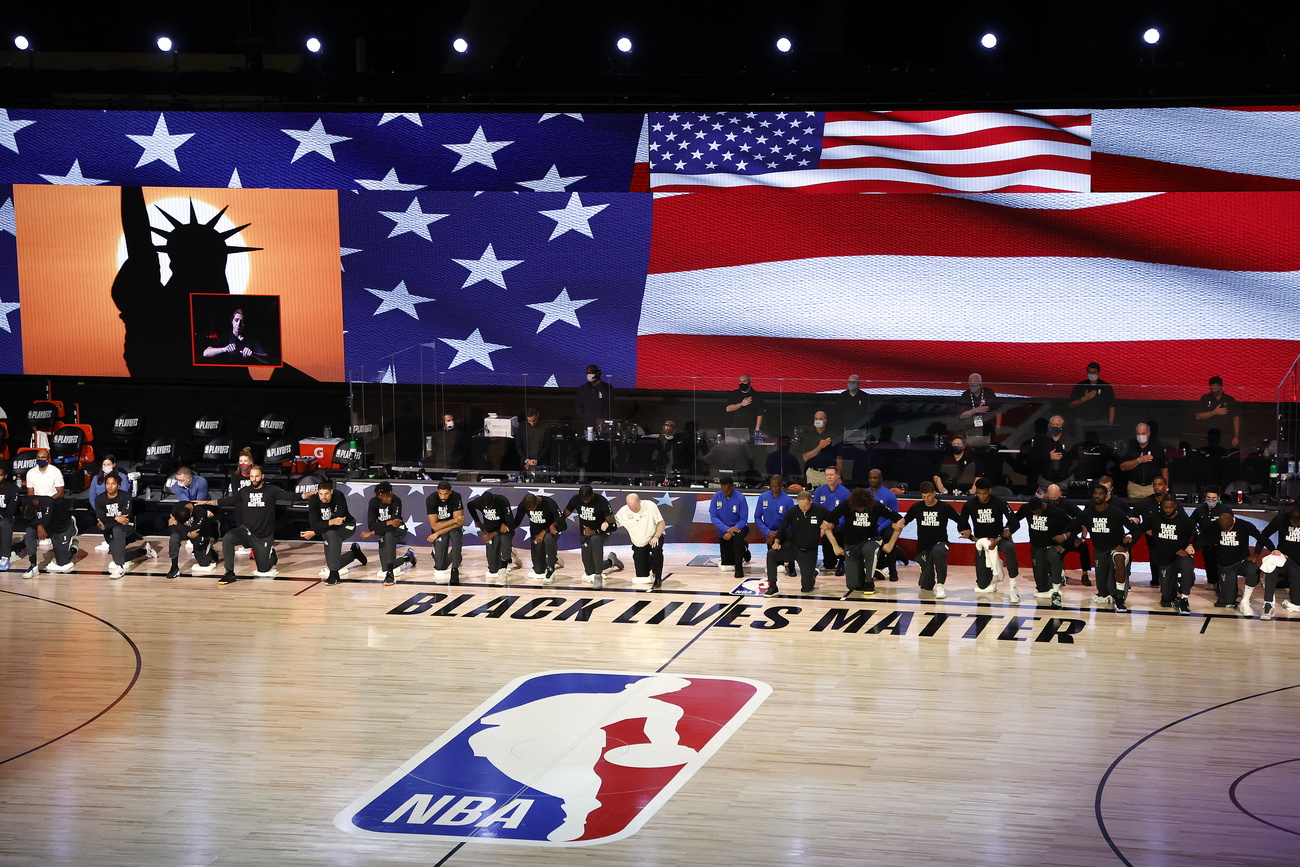 Campo da basket con logo NBA, sul bordo scritta Black Lives Matter e giocatori inginocchiati in rango