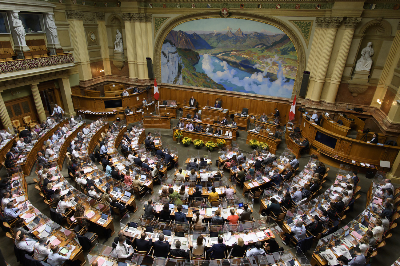 Veduta aerea di una sala parlamentare con grosso dipinto a tema alpino dietro lo scranno del presidente